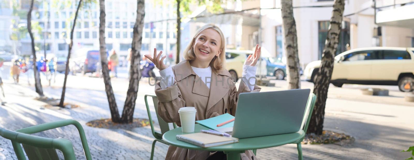 Image of young modern woman, chatting with someone online, having video call via laptop, sitting in outdoor cafe, drinking coffee, gesturing and smiling.