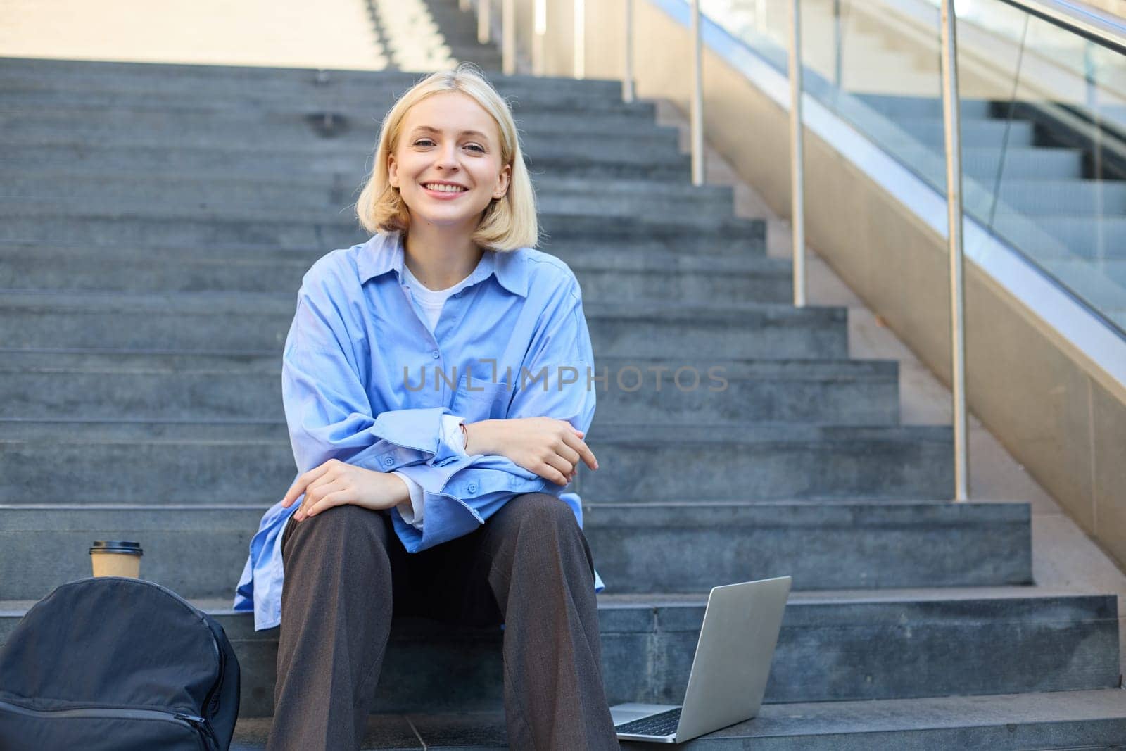 Close up portrait of young cheerful woman, looking confident at camera, sitting on street, urban stairs with laptop, backpack and cup of coffee.