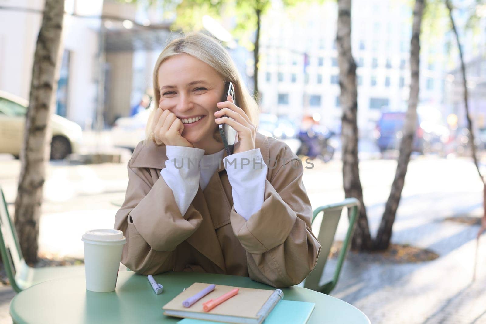 Close up of cheerful blond girl, woman talking on mobile phone, sitting in city centre, outdoor cafe, taking break from work or studying, chatting on telephone by Benzoix