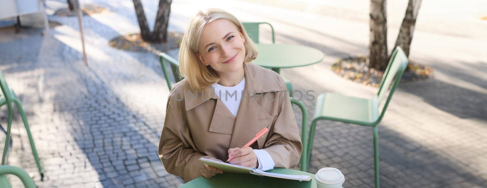 Image of young creative student, woman sitting in outdoor cafe, drawing in notebook, creating sketches in coffee shop, working on art project, smiling at camera.