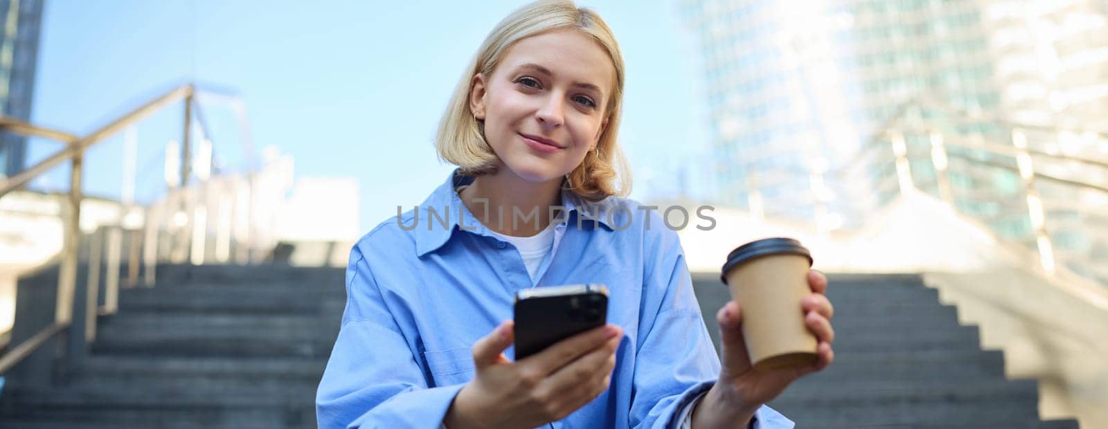 Close up portrait of young smiling woman with cup of coffee, drinking and sitting on stairs in city, holding smartphone in hand by Benzoix