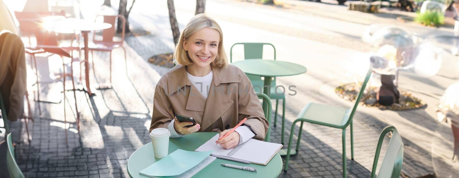 Portrait of smiling young woman using mobile phone while doing homework, working on project, sitting outdoors in cafe, drinking coffee and studying, holding smartphone by Benzoix
