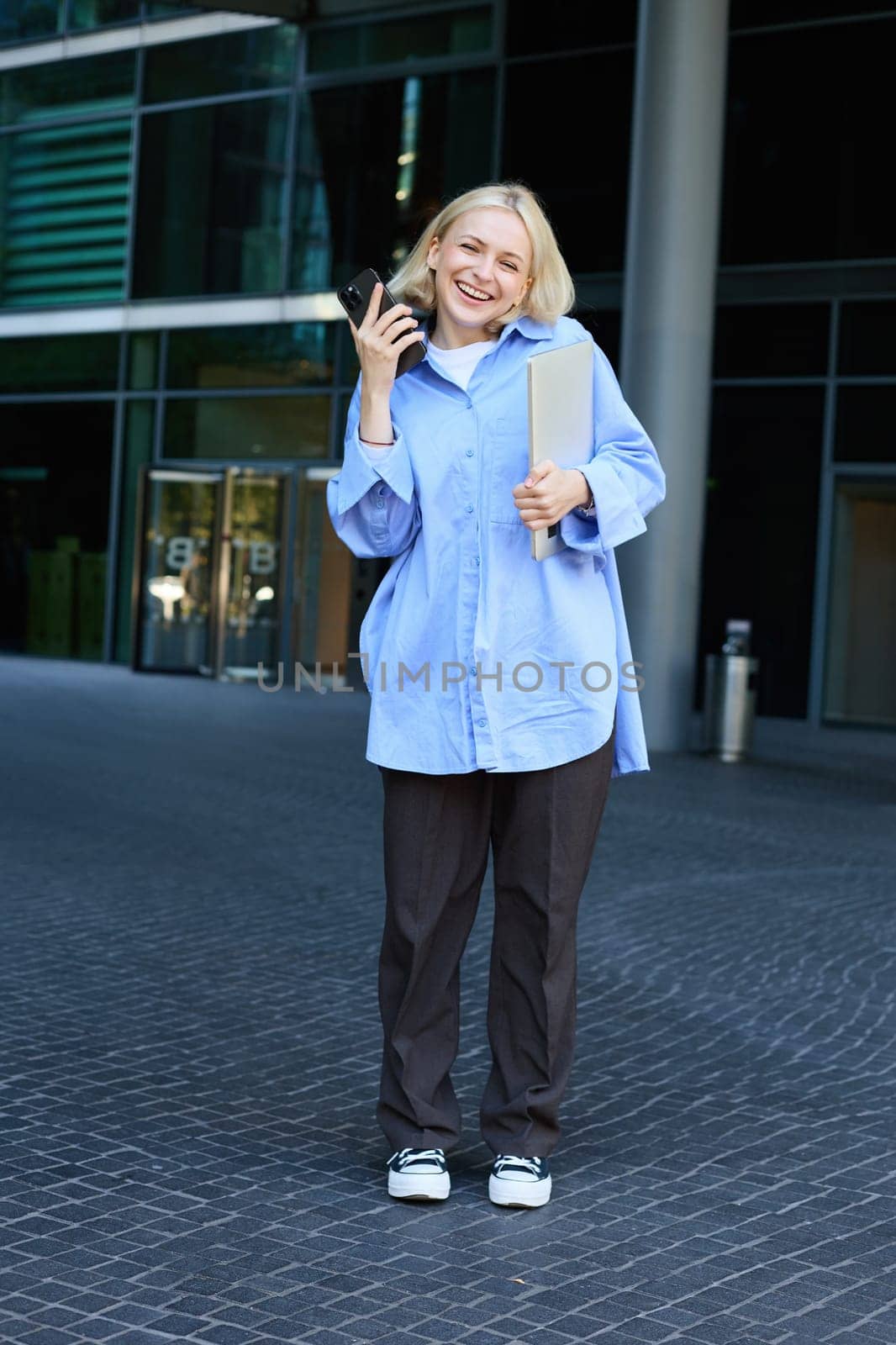 Vertical shot of modern, stylish young blond woman smiling, posing in trousers and blue collar shirt, holding smartphone and laughing, posing near campus building, has laptop in hand by Benzoix