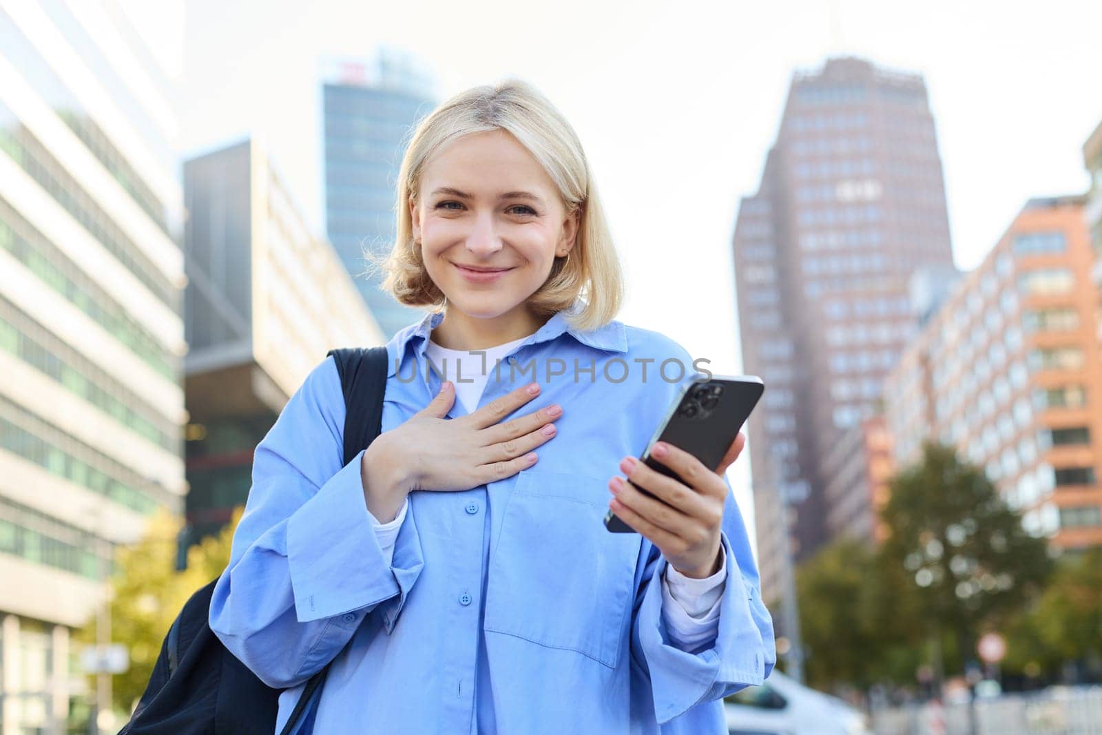 Image of young professional, office manager woman with backpack and smartphone, posing on streets of busy city, holding hand on chest, smiling and looking excited at camera.