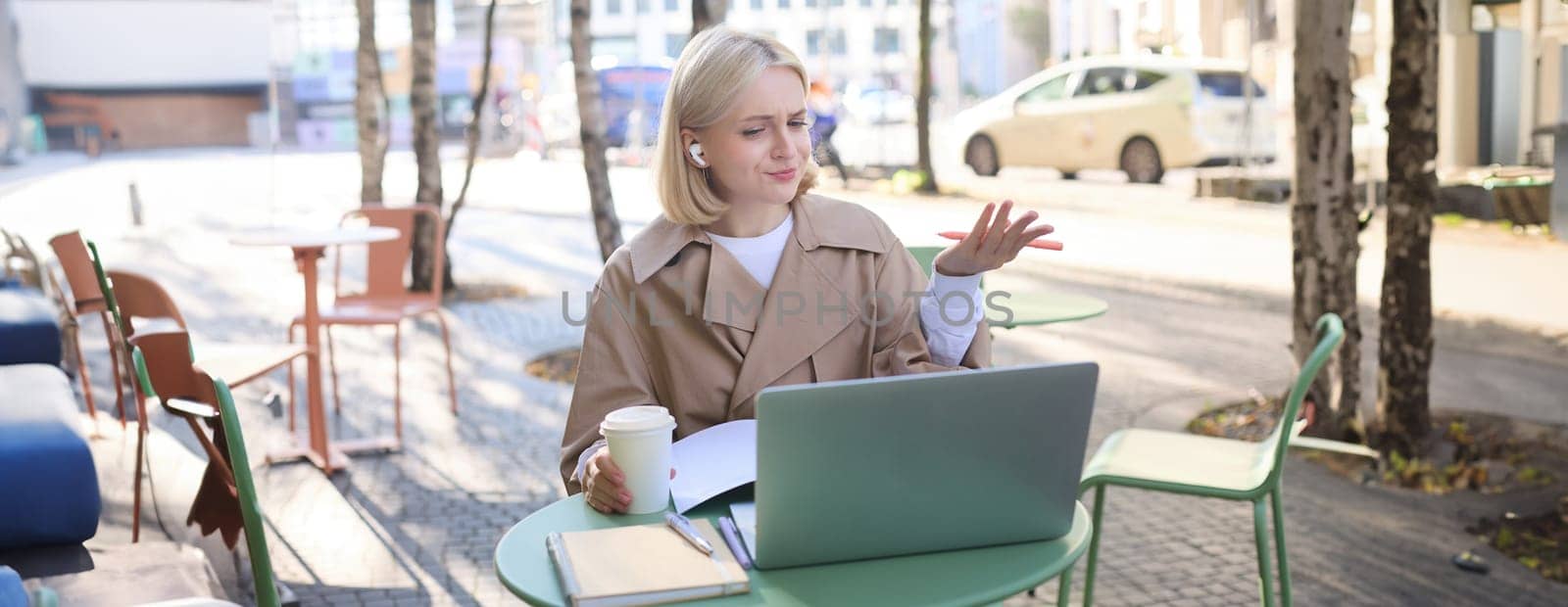 Image of young woman with puzzled face, looking at laptop in wireless headphones, confused while listening to lecture or online speaker, attend web course while sitting in outdoor cafe.