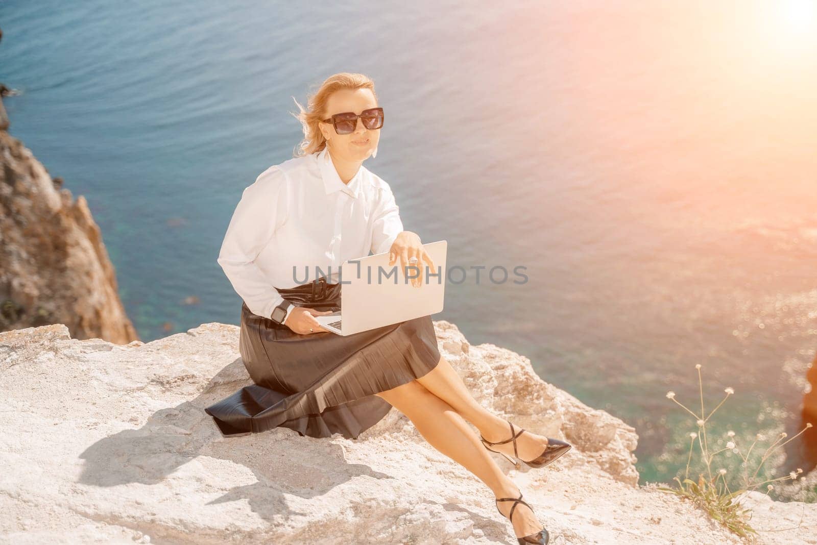 Business woman on nature in white shirt and black skirt. She works with an iPad in the open air with a beautiful view of the sea. The concept of remote work