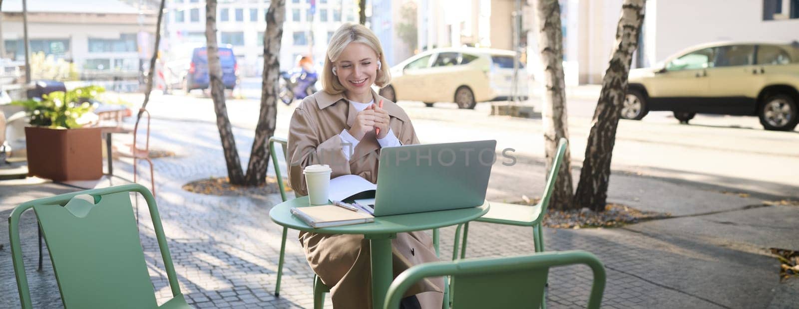 Cheerful, beautiful smiling woman sitting at table in street cafe, working outdoors on sunny warm day, using laptop, wearing wireless headphones, watching online course.
