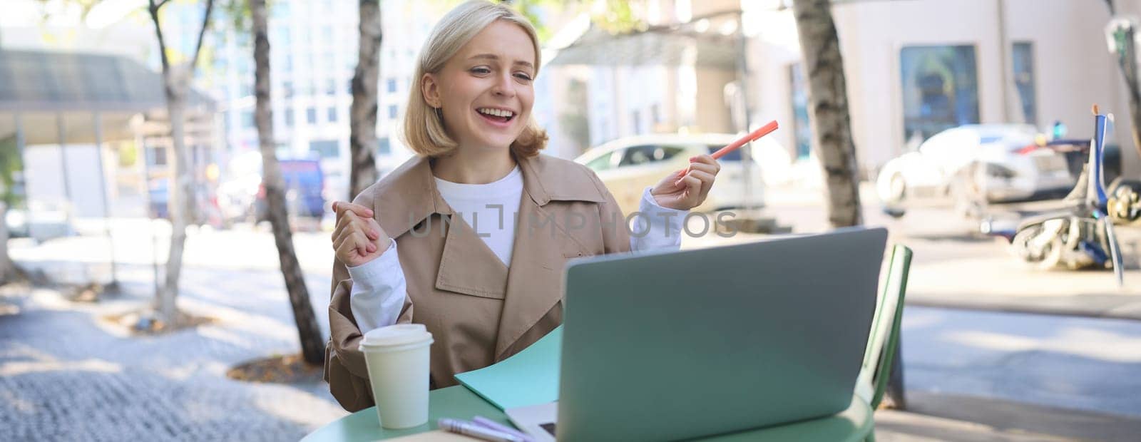 Portrait of young blond woman, modern female model sitting in outdoor cafe, drinking coffee, connects to online video chat via laptop, talking to coworkers remotely, gesturing and explaining smth by Benzoix