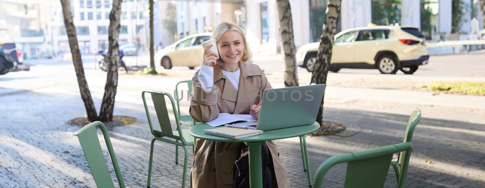 Portrait of young smiling blond woman, working on laptop, sitting in outdoor cafe on street, drinking coffee. Lifestyle and people concept