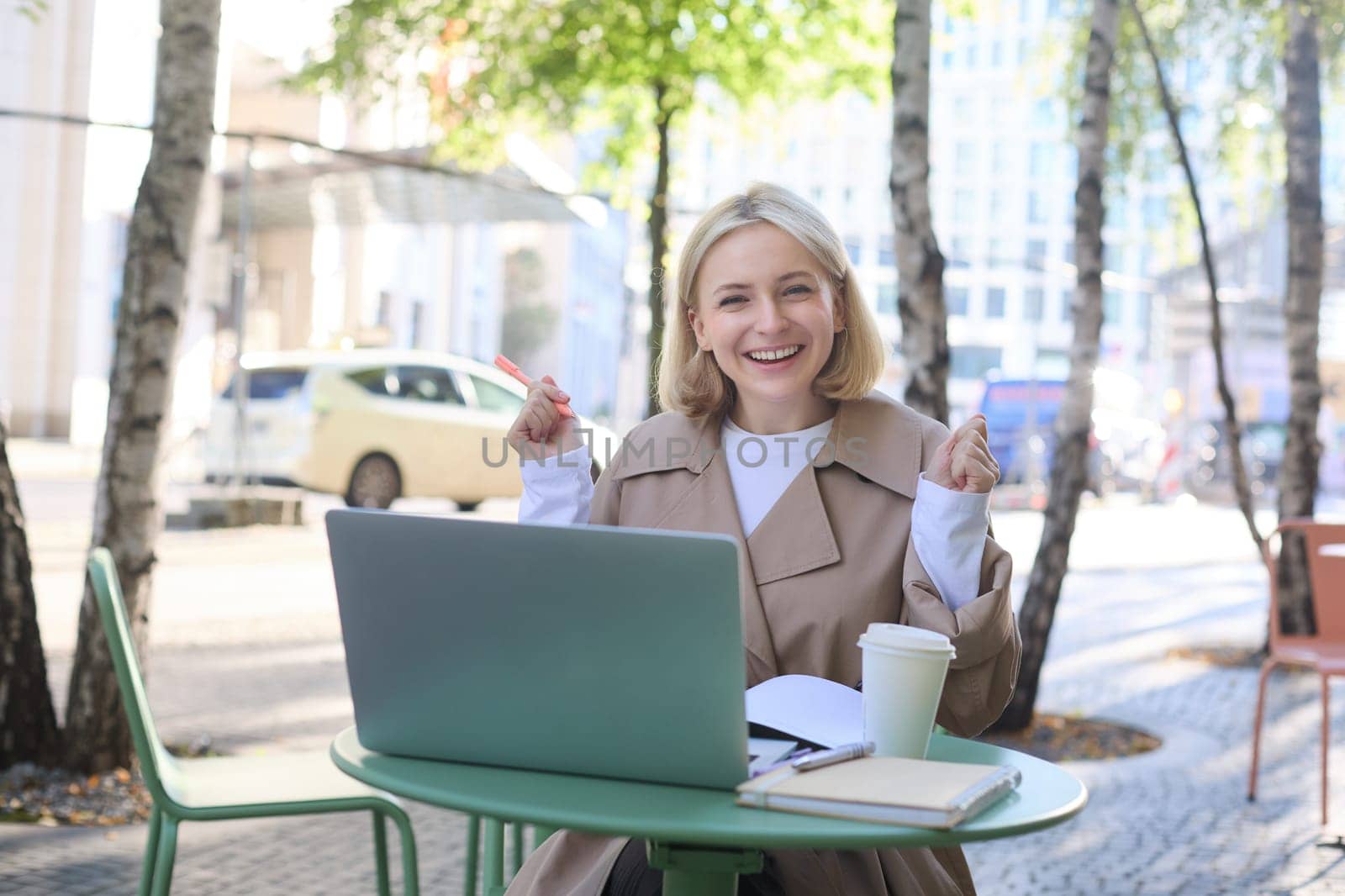Lifestyle portrait of young surprised woman, looking at laptop with amazed face, sitting in coffee shop with excited face expression, spending time outdoors on sunny day.