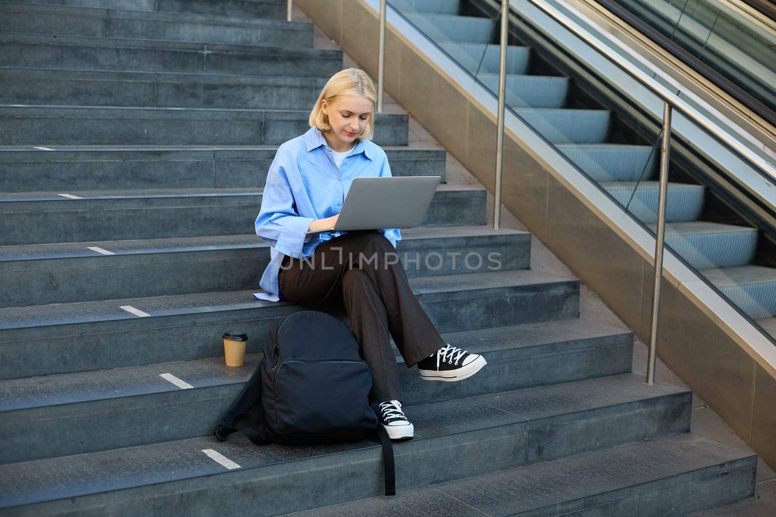 Portrait of woman with laptop, found wifi spot, sitting on stairs outdoors, drinking coffee, working on computer.