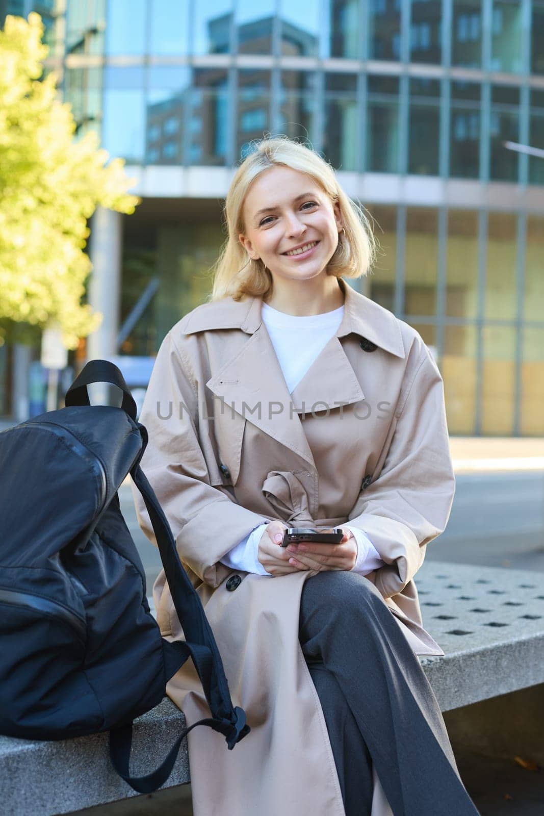 Vertical shot of cute blond woman in trench coat, sitting on street bench with smartphone, chatting with friend, waiting for someone outside, smiling and looking at camera.