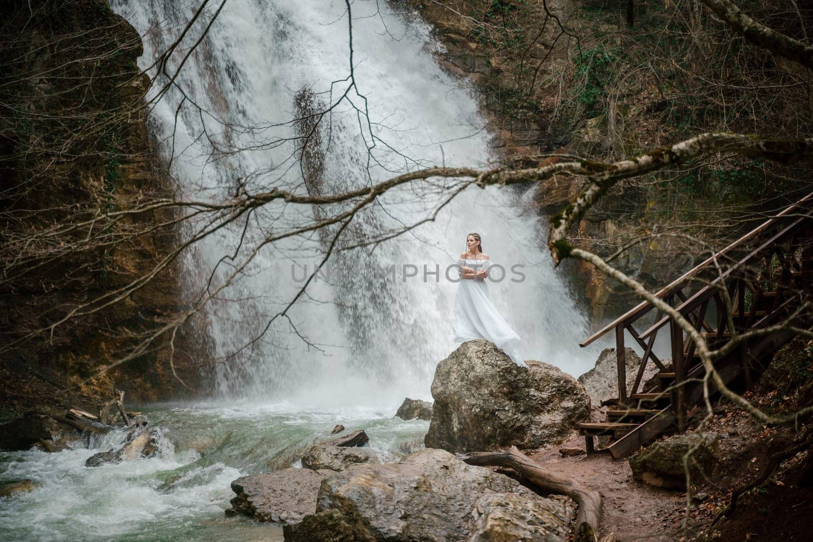 Happy woman in a white dress stands on a stone with a waterfall behind.