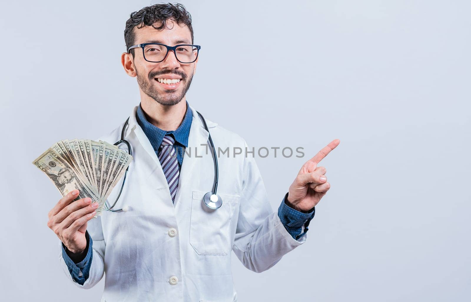 Happy young doctor holding money and pointing to side. Handsome smiling doctor showing money and pointing side. Latin doctor