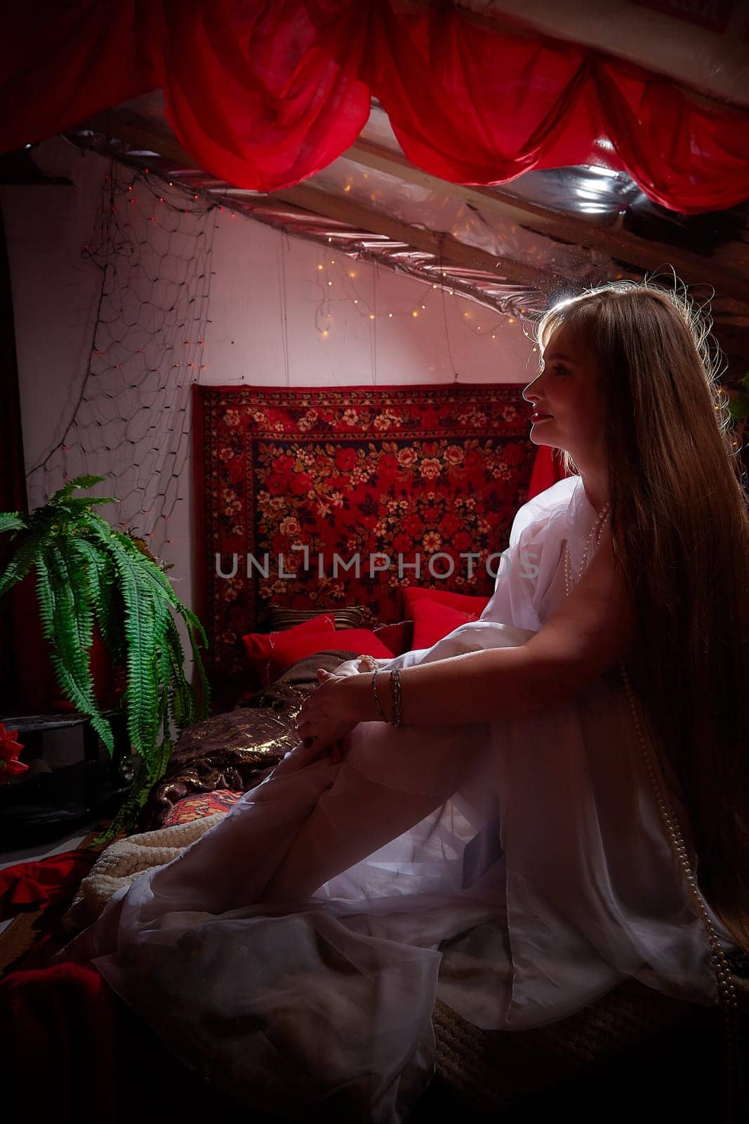 Beautiful European girl looking like Arab woman in red room with rich fabrics and carpets in sultan harem. Photo shoot of an oriental style odalisque. A model poses in sari as indian woman in india