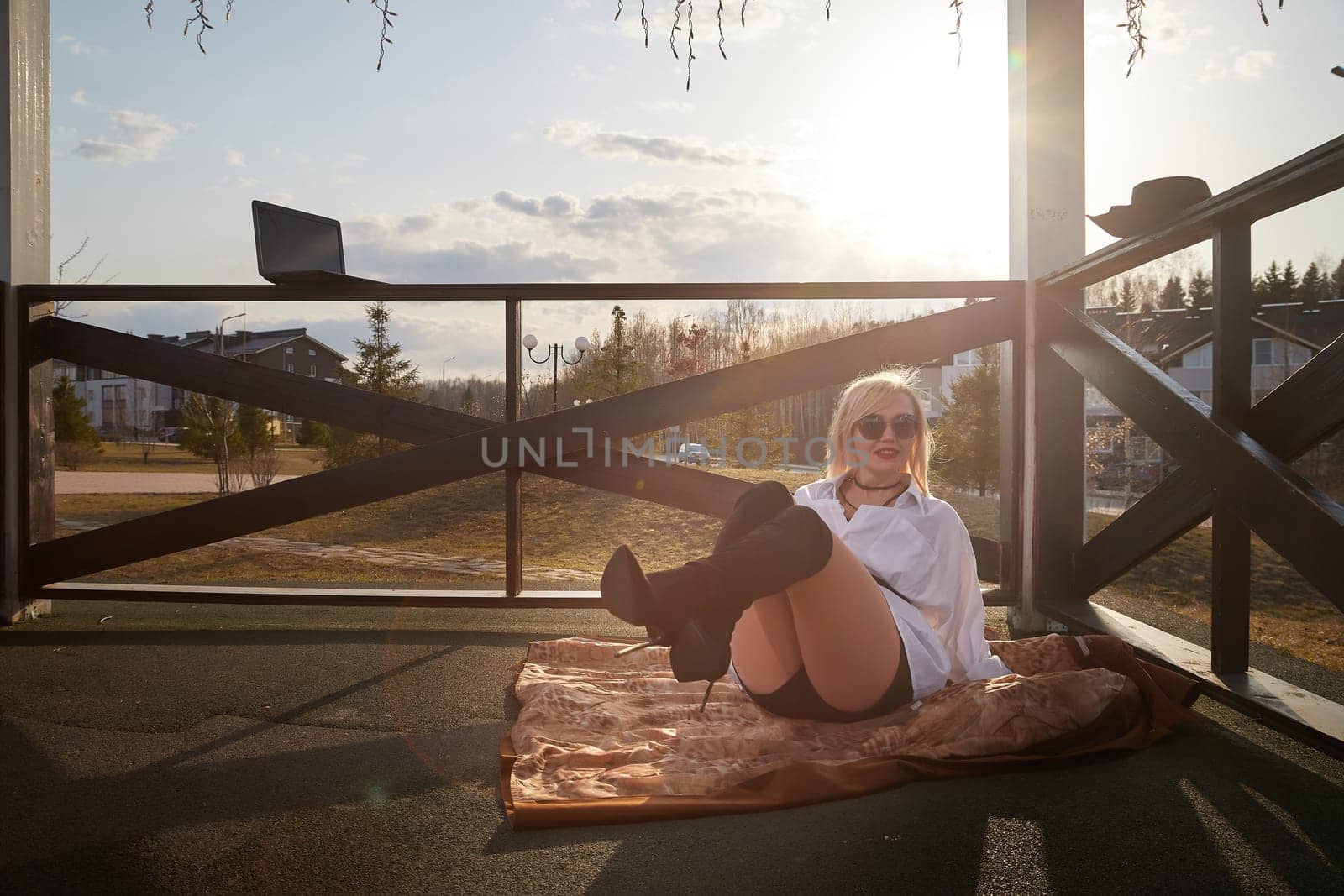 Fashionable beautiful blonde girl in a white shirt and a short skirt is resting in a gazebo with a laptop on sunny autumn, spring day. Lazy and relaxing businesswoman, manager, student in nature