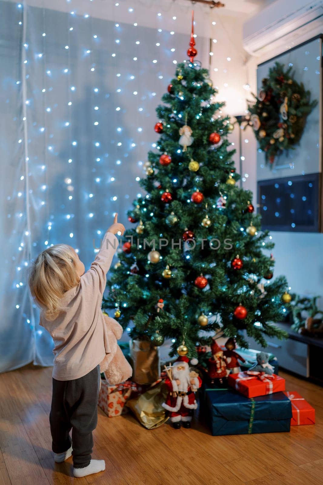 Little girl stands with a teddy bear near a decorated Christmas tree and points to the top. Back view. High quality photo