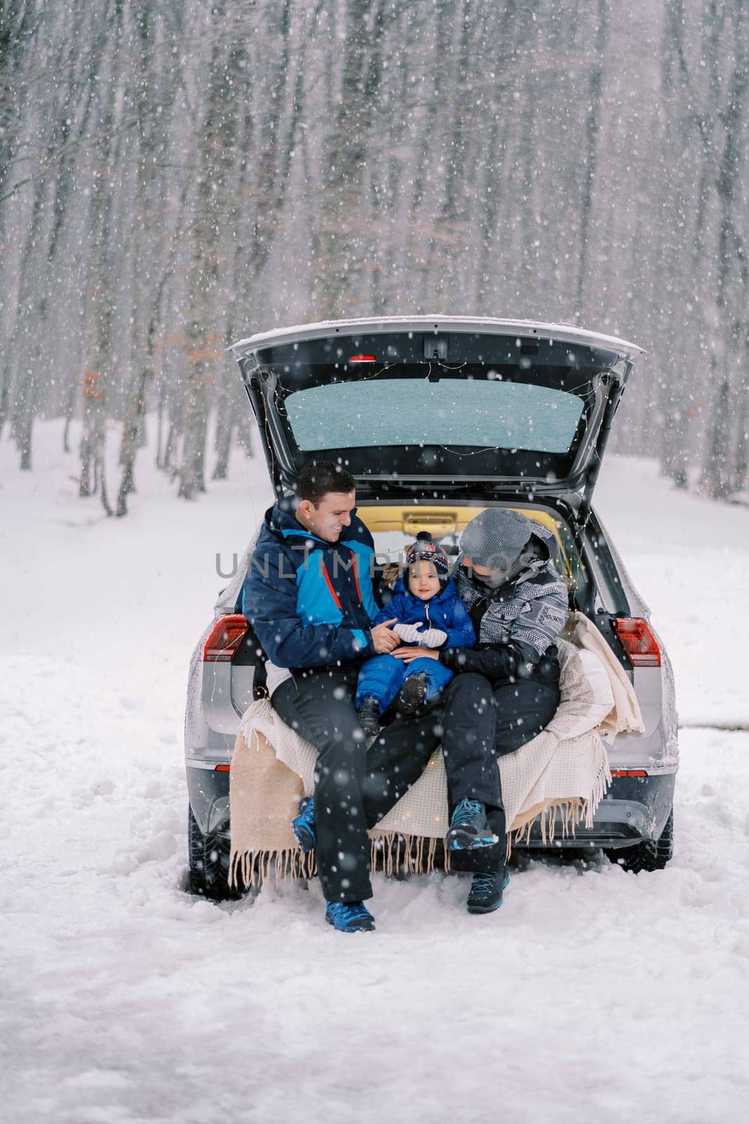 Mom and dad look at a little girl sitting on a blanket in a car trunk in a winter forest. High quality photo