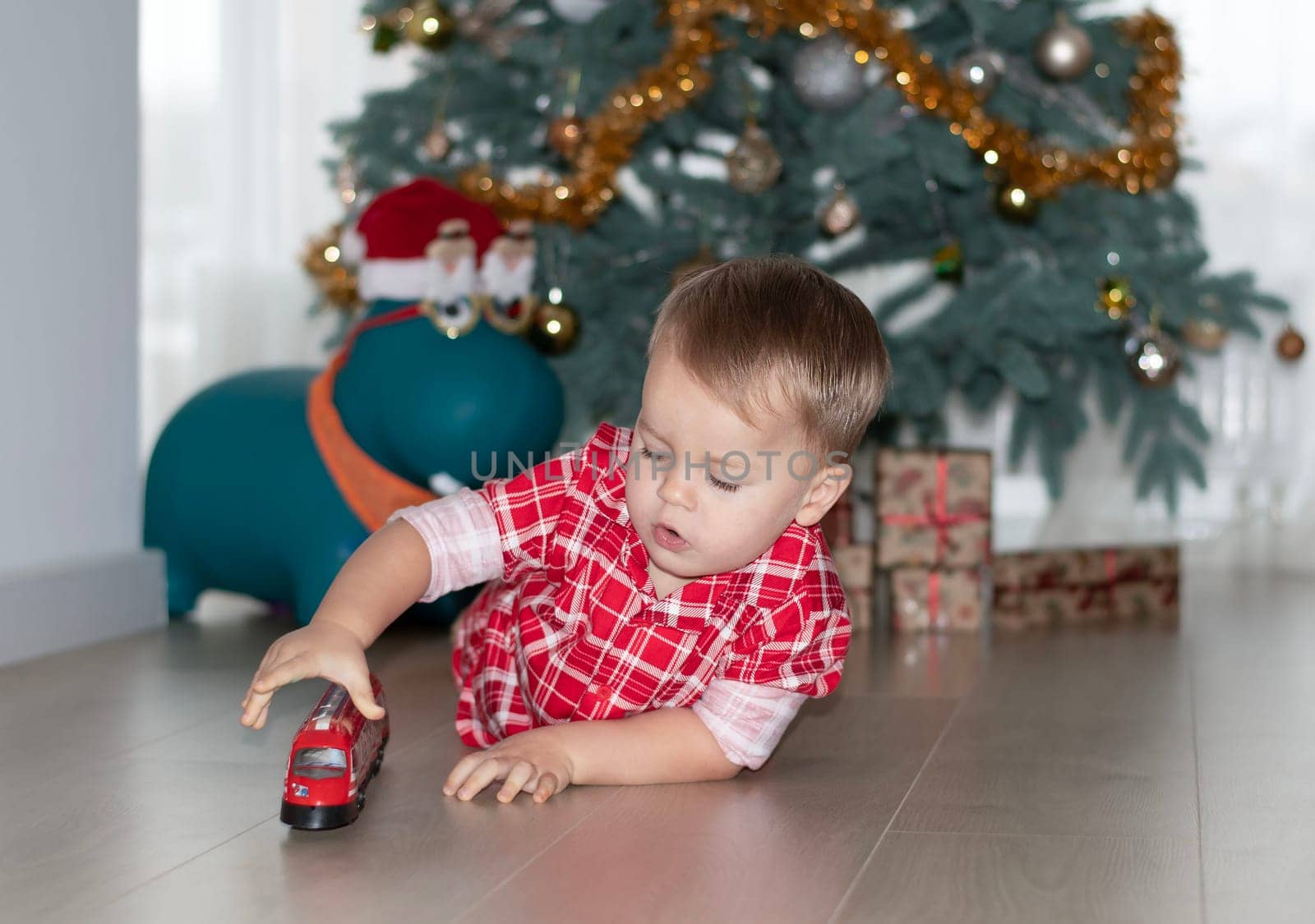 Christmas concept. New Year 2024. A small handsome boy in red checkered pajamas lies on the floor and plays with a red steam locomotive against the background of a decorated Christmas tree with gifts. Close-up.