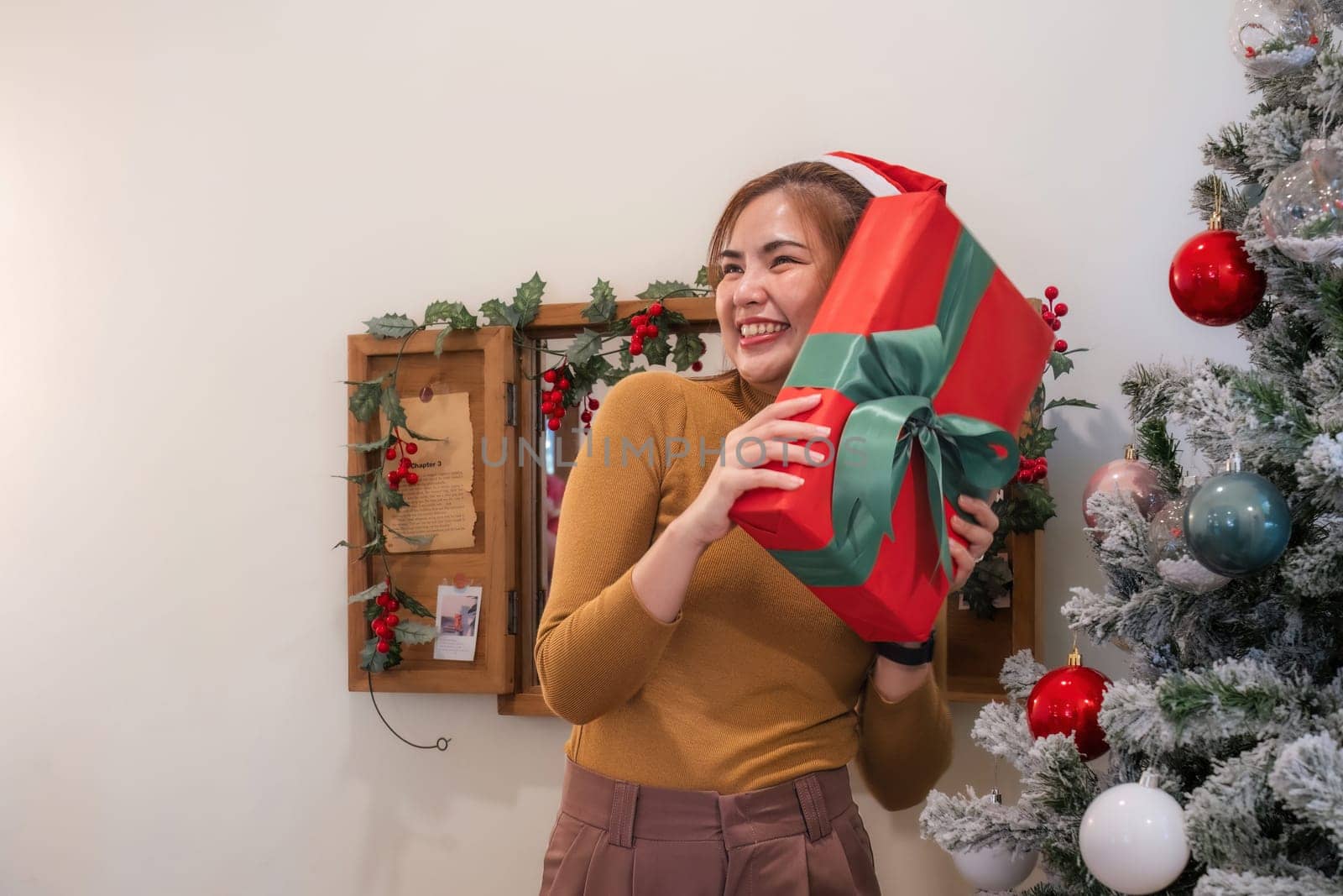 christmas time Adorable young woman with Santa hat and attractive and beautiful gift box for Christmas party in yellow long sleeve shirt near Christmas tree at modern home by wichayada