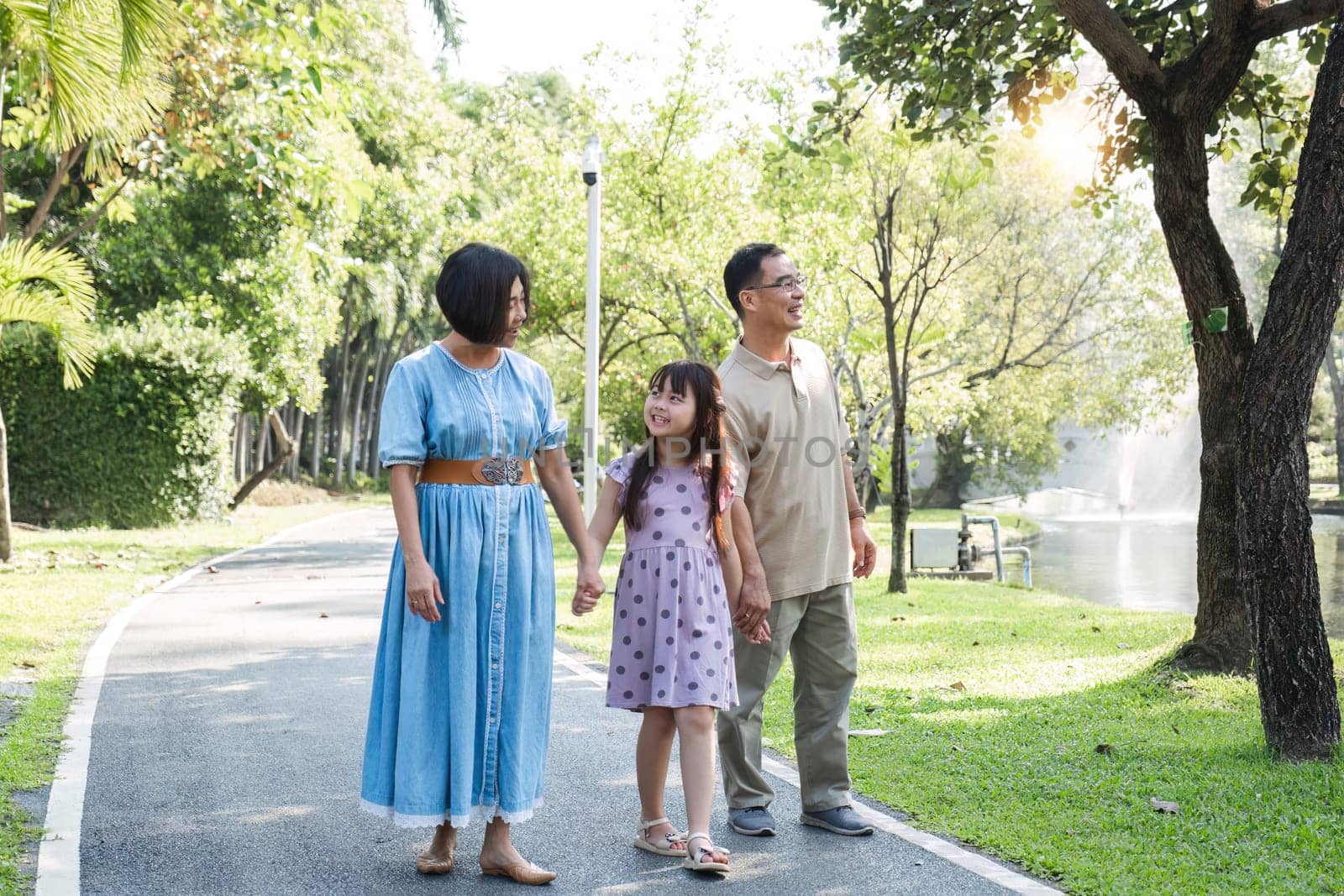 Kind-hearted Asian senior grandparent Warm and happy enjoying a stroll through the park with their adorable little granddaughter. On a clear day together lovely family by wichayada