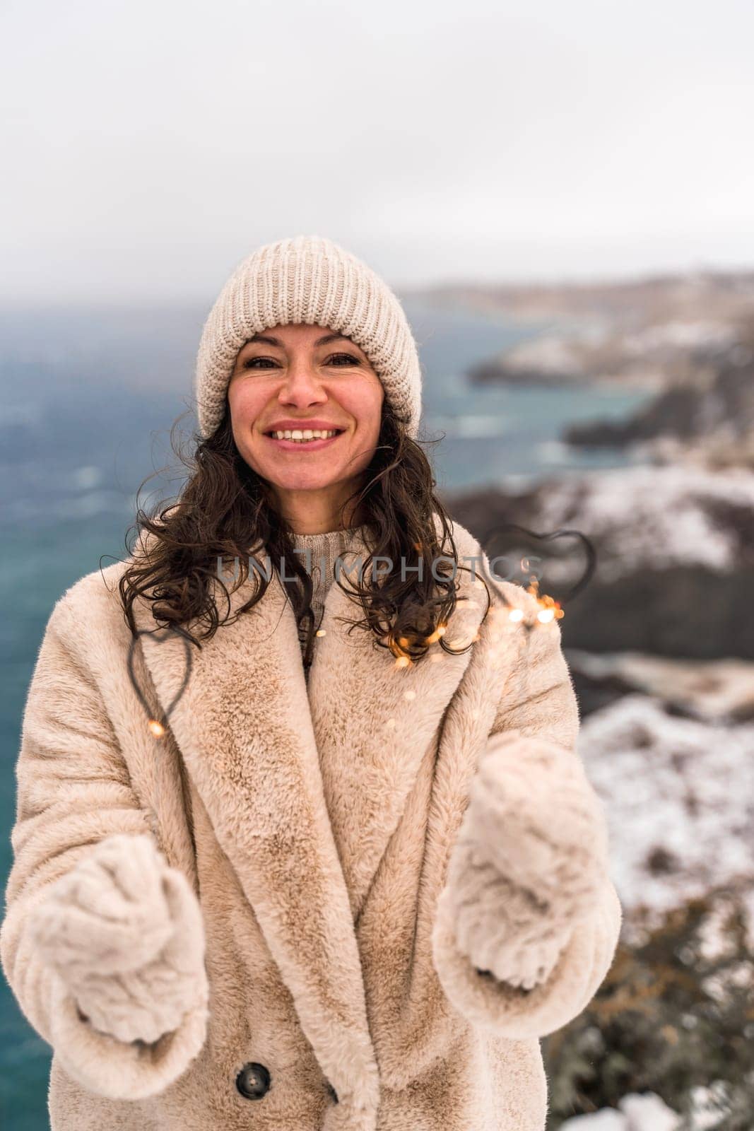 Outdoor winter portrait of elegant happy smiling beige hat, light faux fur coat holding heart sparkler, posing against sea and snow background