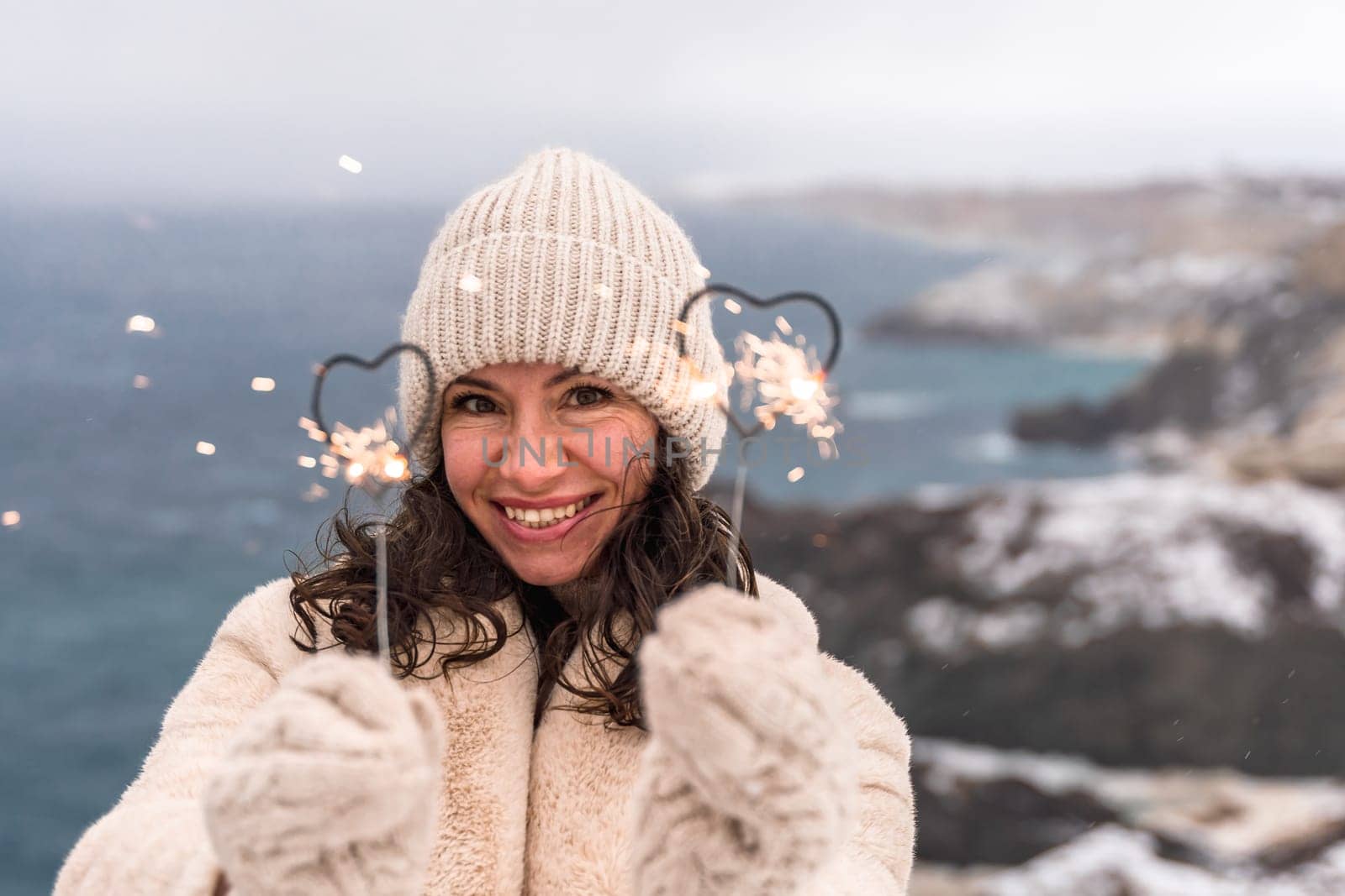 Outdoor winter portrait of elegant happy smiling beige hat, light faux fur coat holding heart sparkler, posing against sea and snow background