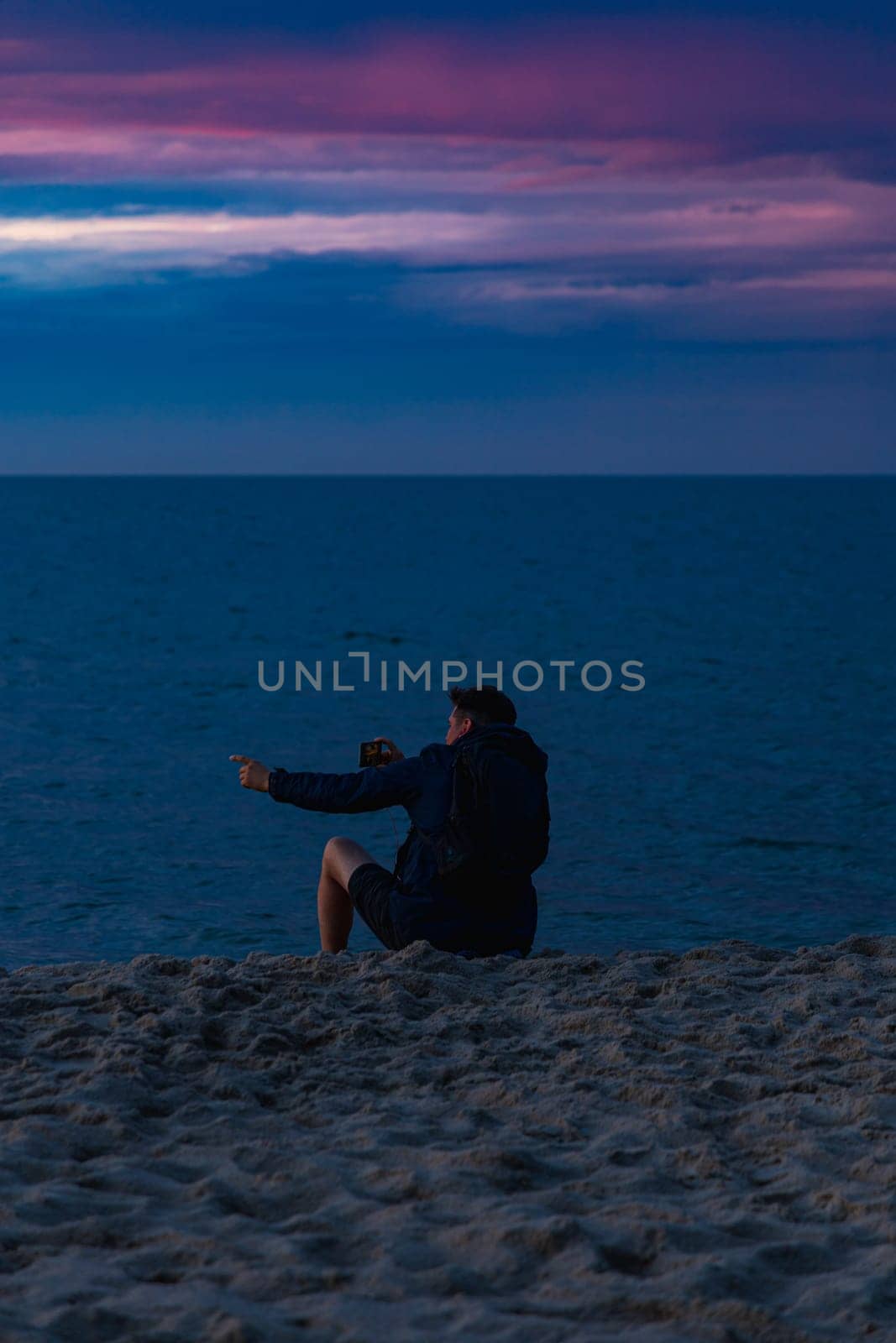 Man who is sitting at the edge of the beach next to water having videocall and showing beautiful cloudy sunset at golden hour at the beach next to dark and cold sea at summer right after storm