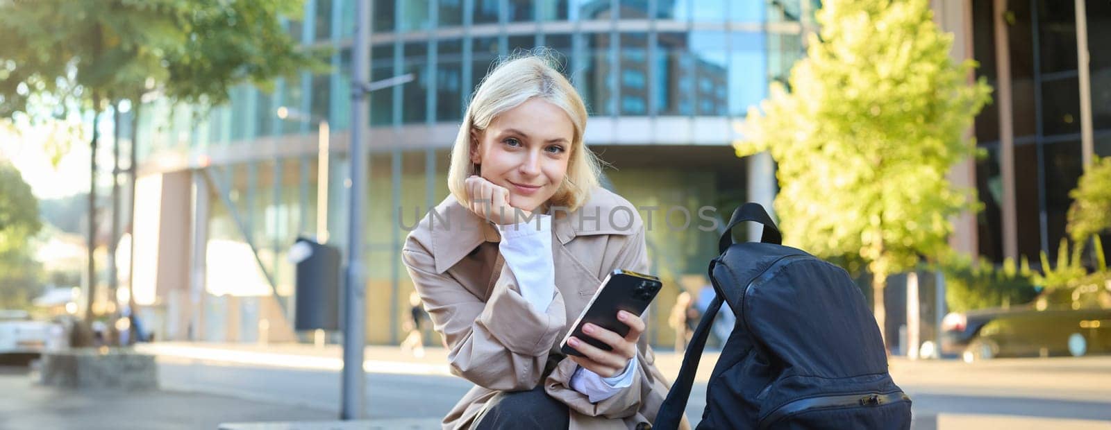 Portrait of young blonde woman, waiting for someone on street bench, sitting with smartphone, using mobile phone app.