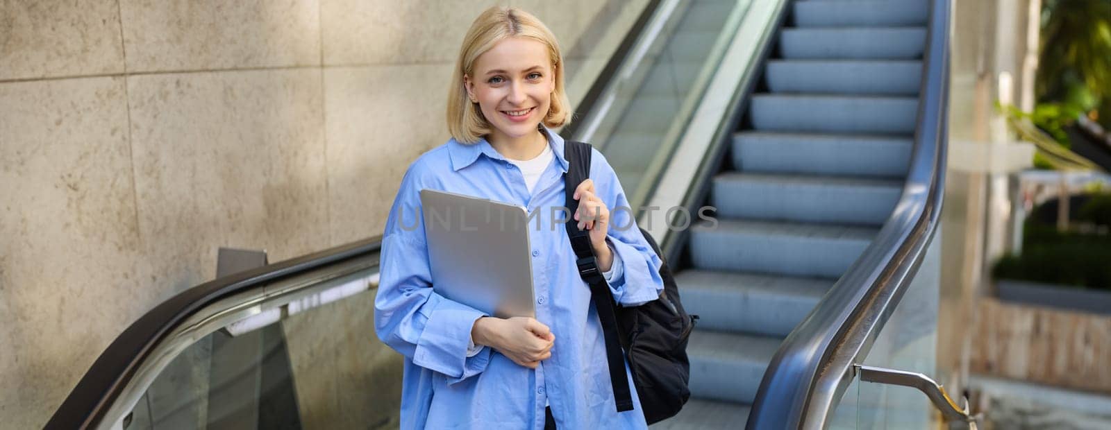 Lifestyle portrait of young blond woman, carries her backpack and laptop, using escalator in city, smiling and looking confident at camera. Education and people concept