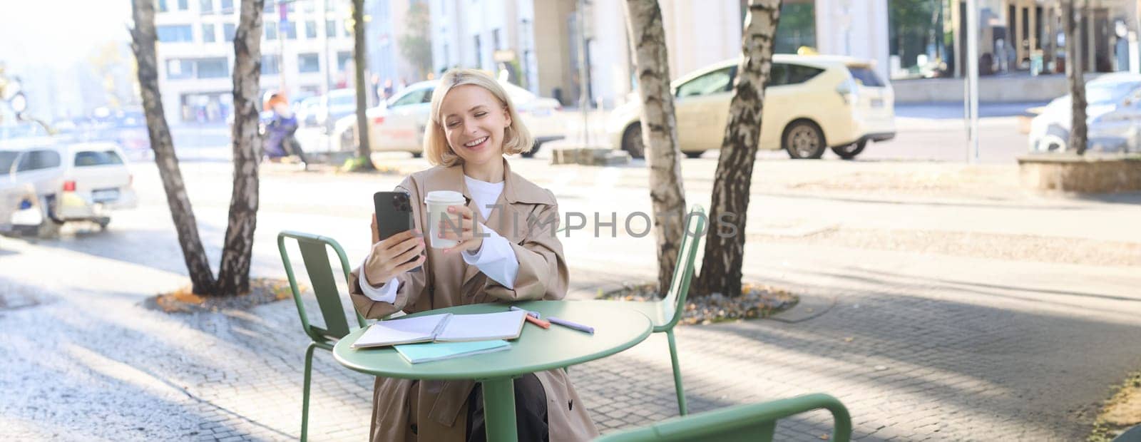 Lifestyle portrait of young woman has video conversation, chatting on smartphone app, showing takeaway cup while sitting outside in coffee shop.