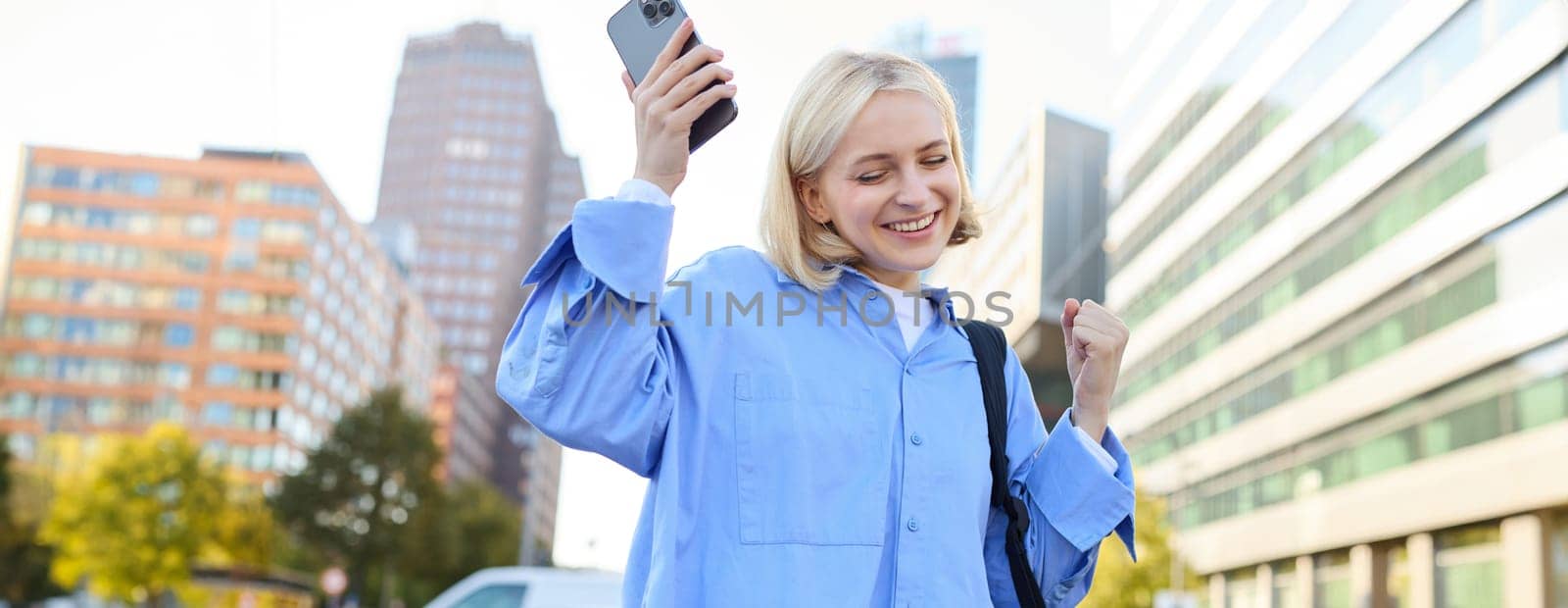 Portrait of happy, enthusiastic young woman, holding backpack and mobile phone, standing on street and rejoicing, triumphing, making fist pump gestures, dancing from happiness and joy.