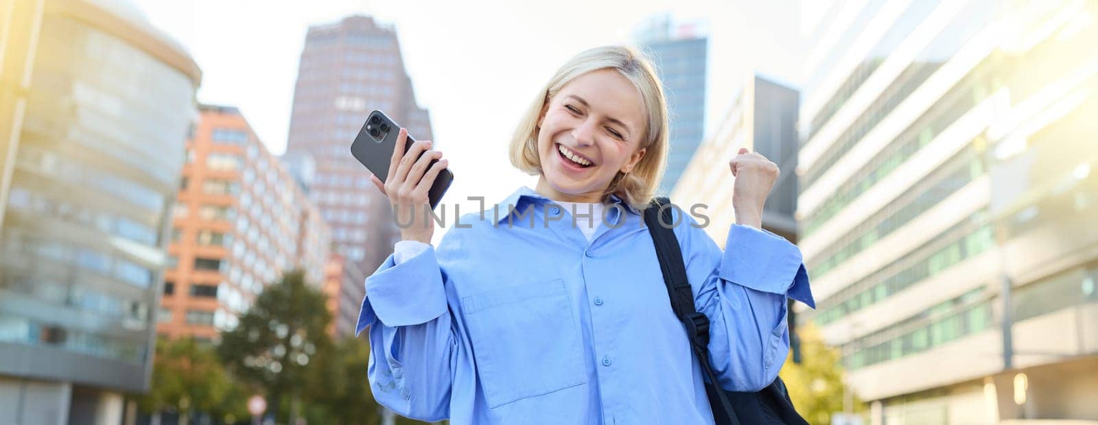 Portrait of happy, enthusiastic young woman, holding backpack and mobile phone, standing on street and rejoicing, triumphing, making fist pump gestures, dancing from happiness and joy.