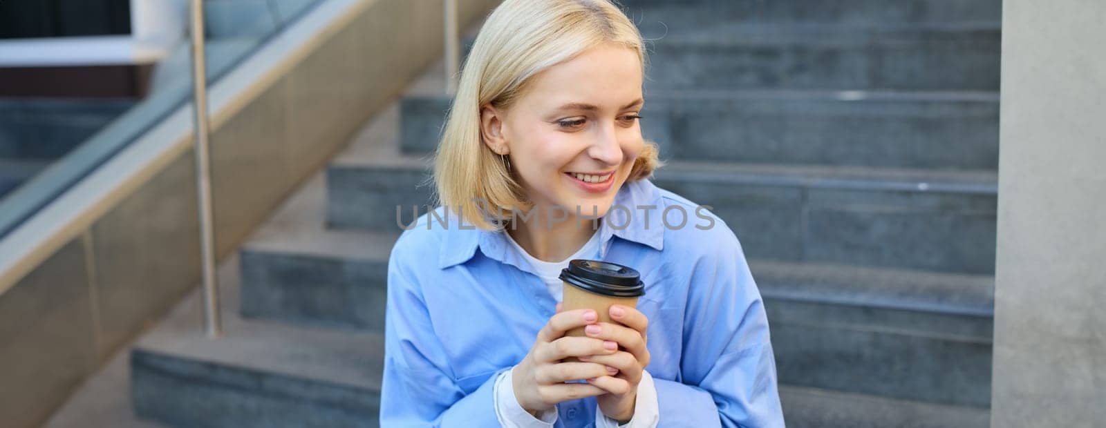 Cute young female student, woman with cup of coffee in hands, resting on stairs in city centre, sitting and drinking cappuccino, smiling happily.