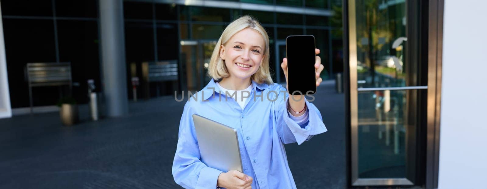 Portrait of stylish, modern young office worker, woman showing her smartphone screen, mobile phone app on display, posing near campus or office building, holding laptop.