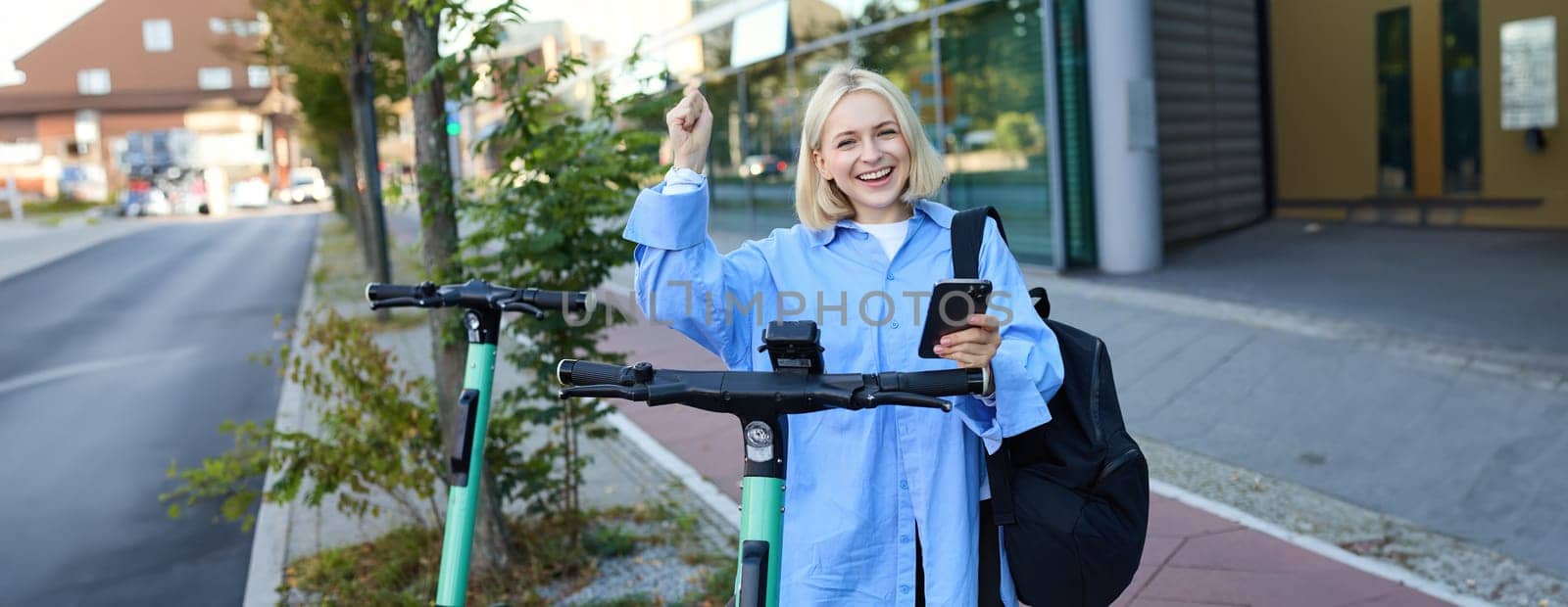 Portrait of young smiling female model, renting an electric scooter, using mobile phone to scan QR code with smartphone app, riding home from university, standing outdoors on street.