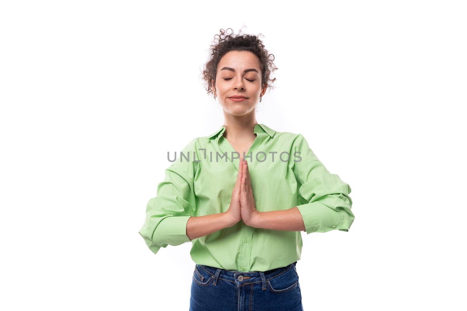 young brunette curly slender female model dressed in a light green shirt meditates with folded arms.