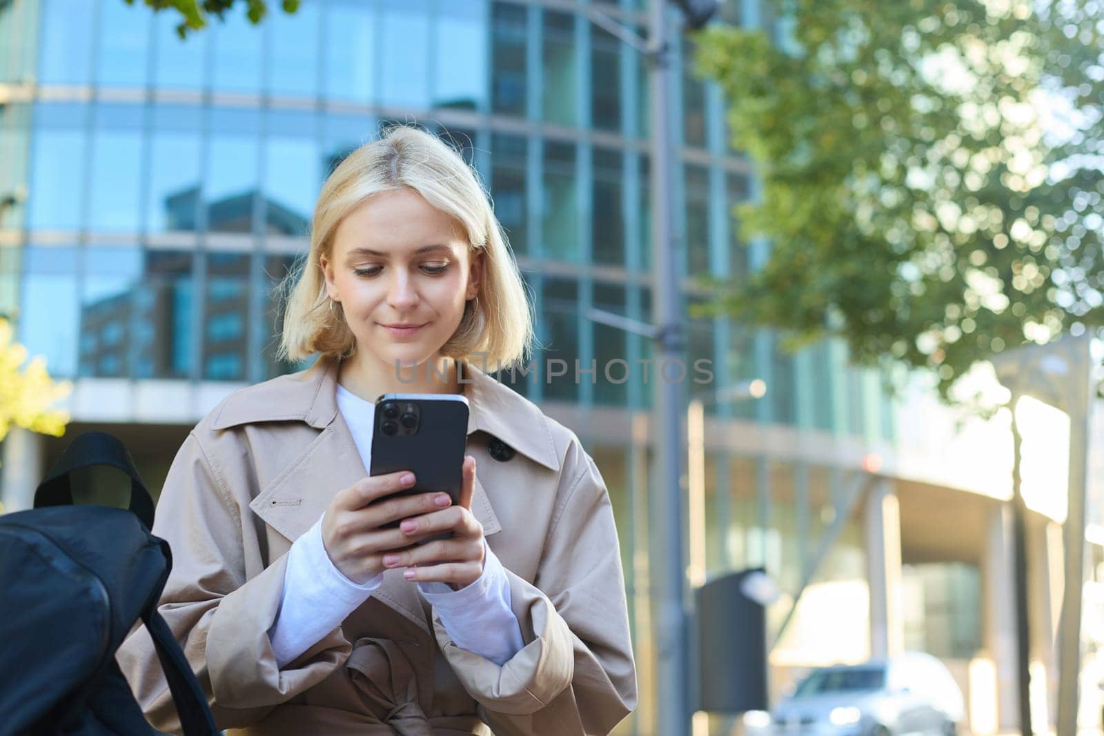 Portrait of young blonde woman, waiting for someone on street bench, sitting with smartphone, using mobile phone app.