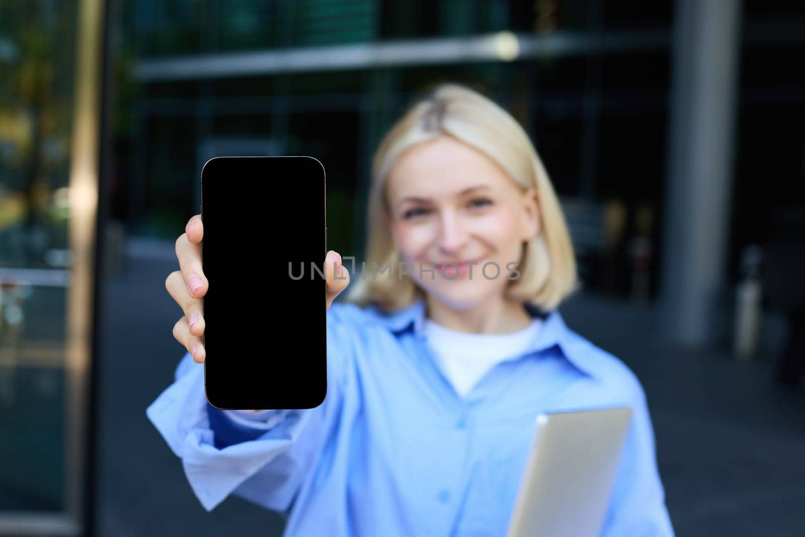 Portrait of beautiful young woman, student posing on street near campus, holding laptop, showing mobile phone screen, demonstrating app or promo on smartphone and smiling by Benzoix