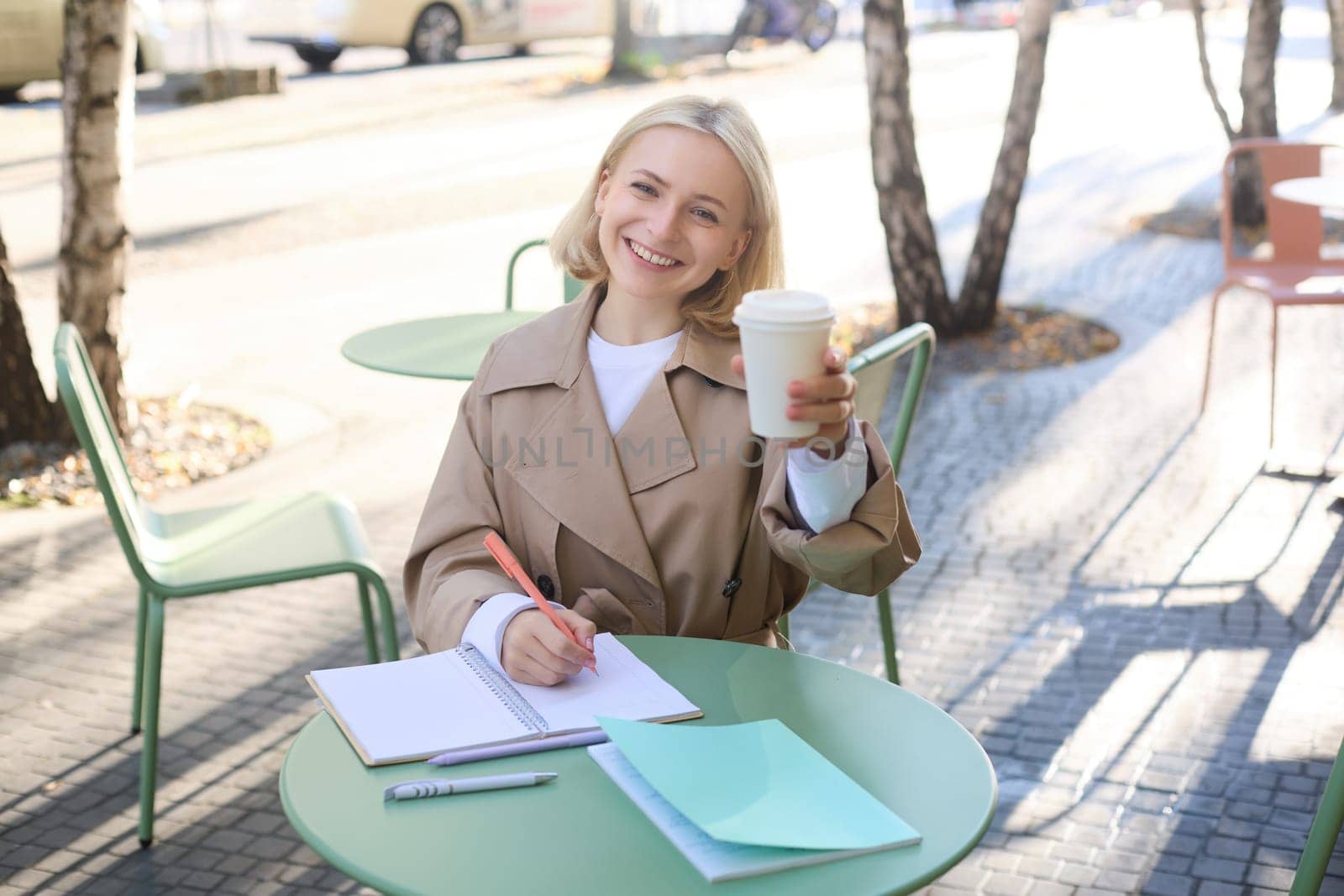 Smiling young blond woman, enjoying her coffee, drinking takeaway and sitting in street cafe, working on project, student doing homework, writing something in notebook, looking happy at camera.
