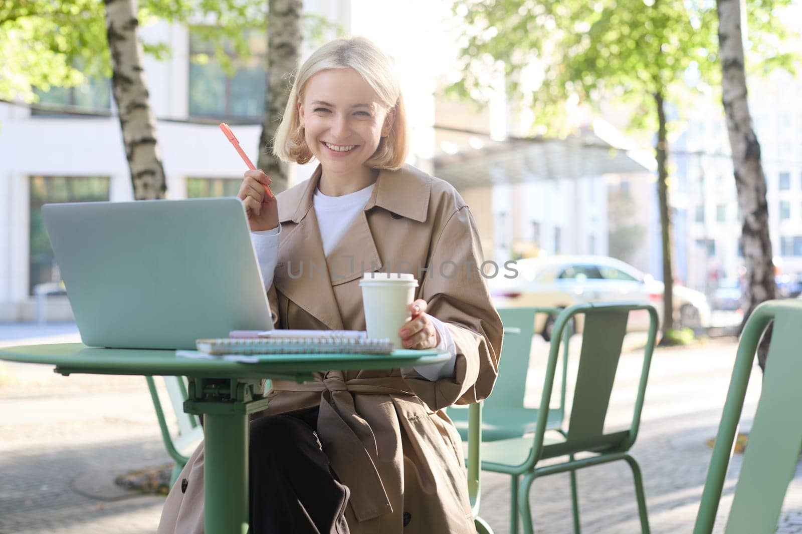 Portrait of young smiling blond woman, working on laptop, sitting in outdoor cafe on street, drinking coffee by Benzoix