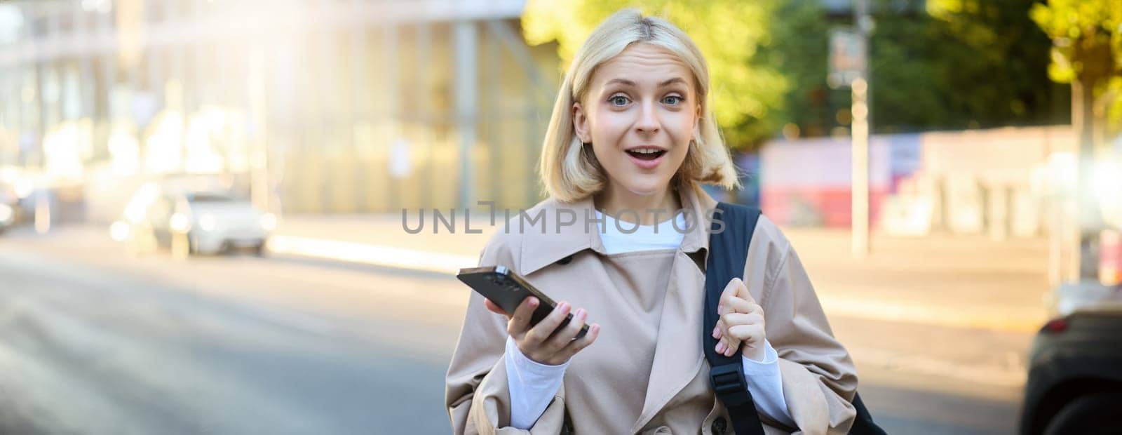Portrait of young blond woman with backpack, holding mobile phone, standing on street on sunny day, looking surprised and amazed at camera, expresses interest, hear great news.