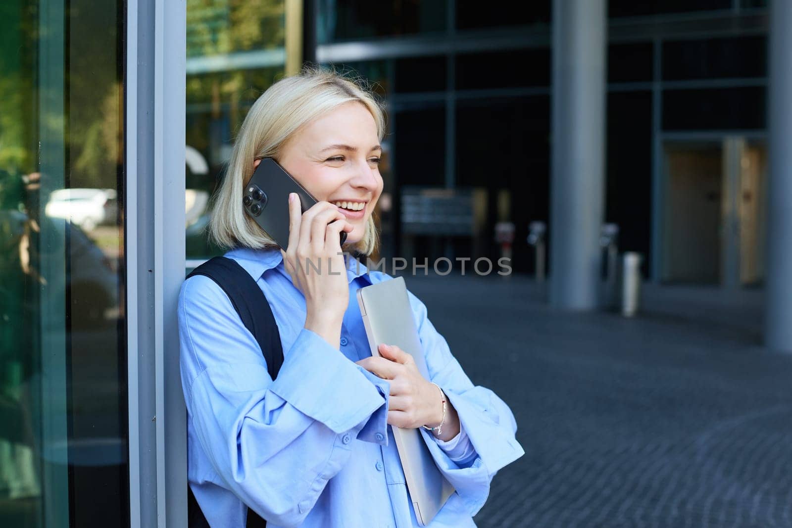 Happy female model, blond woman talking on mobile phone, chatting with someone, standing near office building with laptop and smartphone, smiling by Benzoix