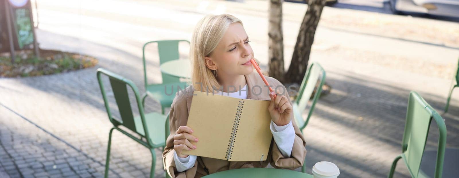 Image of young woman with thoughtful face, holding notebook and pen, thinking, making sketches, thinking, doing homework, writing in her journal while sitting in coffee shop.