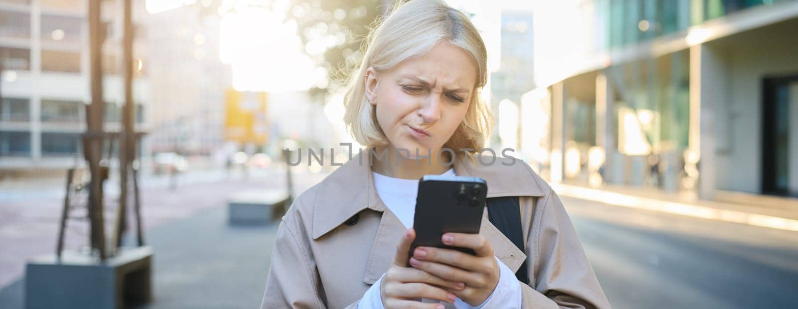 Close up portrait of woman looking sceptical at her mobile phone, standing on street, frowning and standing perplexed, reading something on smartphone screen.