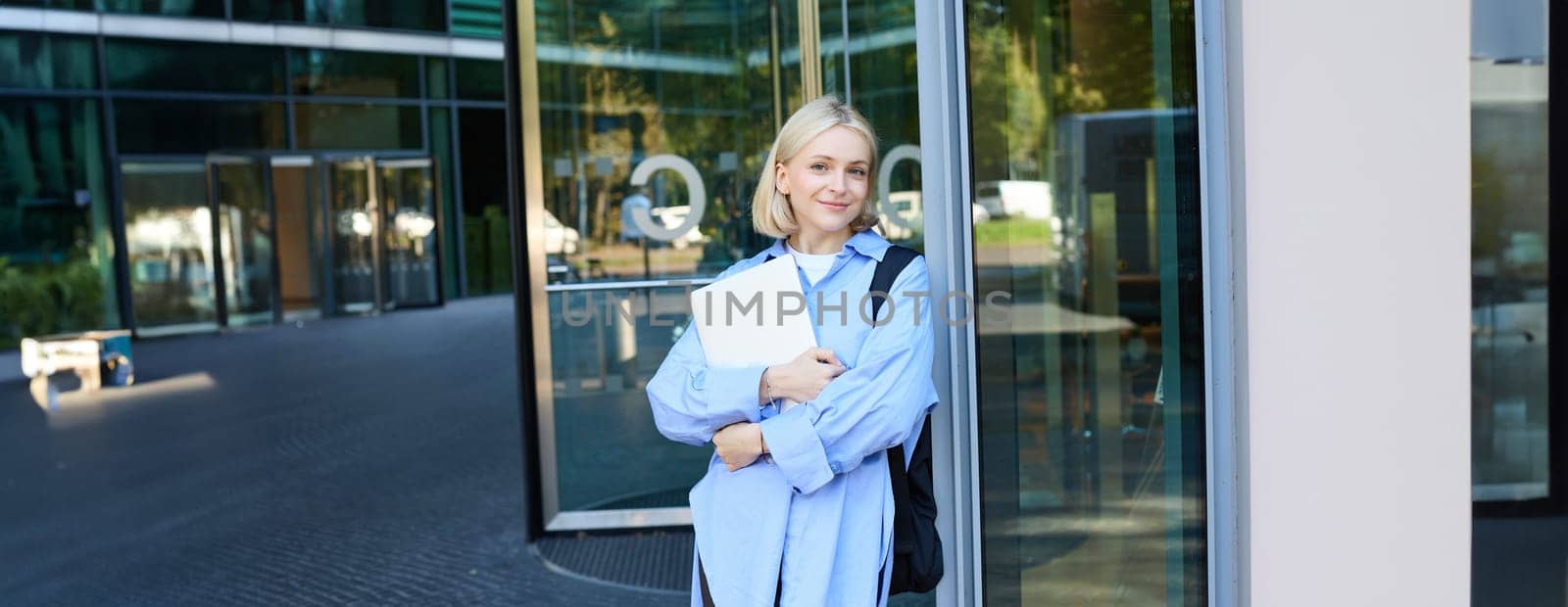 Lifestyle portrait of young woman with laptop, posing near campus, entrance to office building, holding backpack, smiling and looking happy by Benzoix