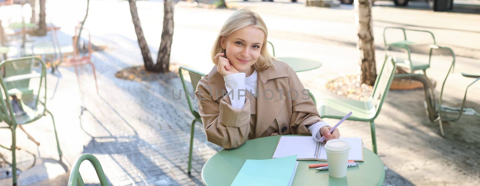 Modern student working on essay in outdoor cafe, doing homework, sitting alone and writing, drinking cup of coffee, smiling at camera.