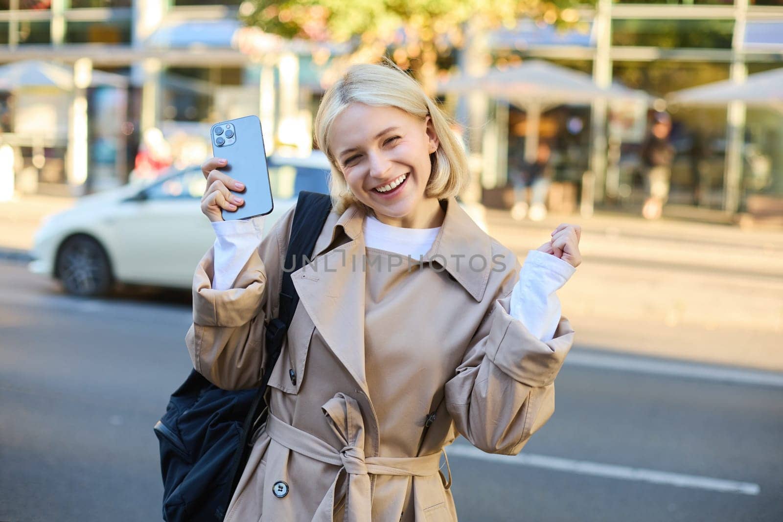 Excited young blond woman with smartphone, standing on street and rejoicing, celebrating victory, triumphing with happy face by Benzoix