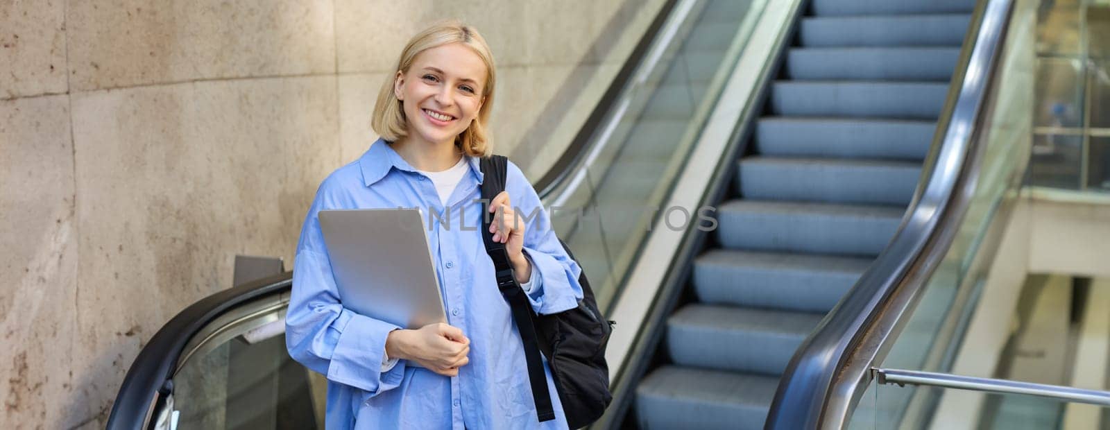 Image of young modern woman, student on escalator, holding laptop and backpack, walking in city.