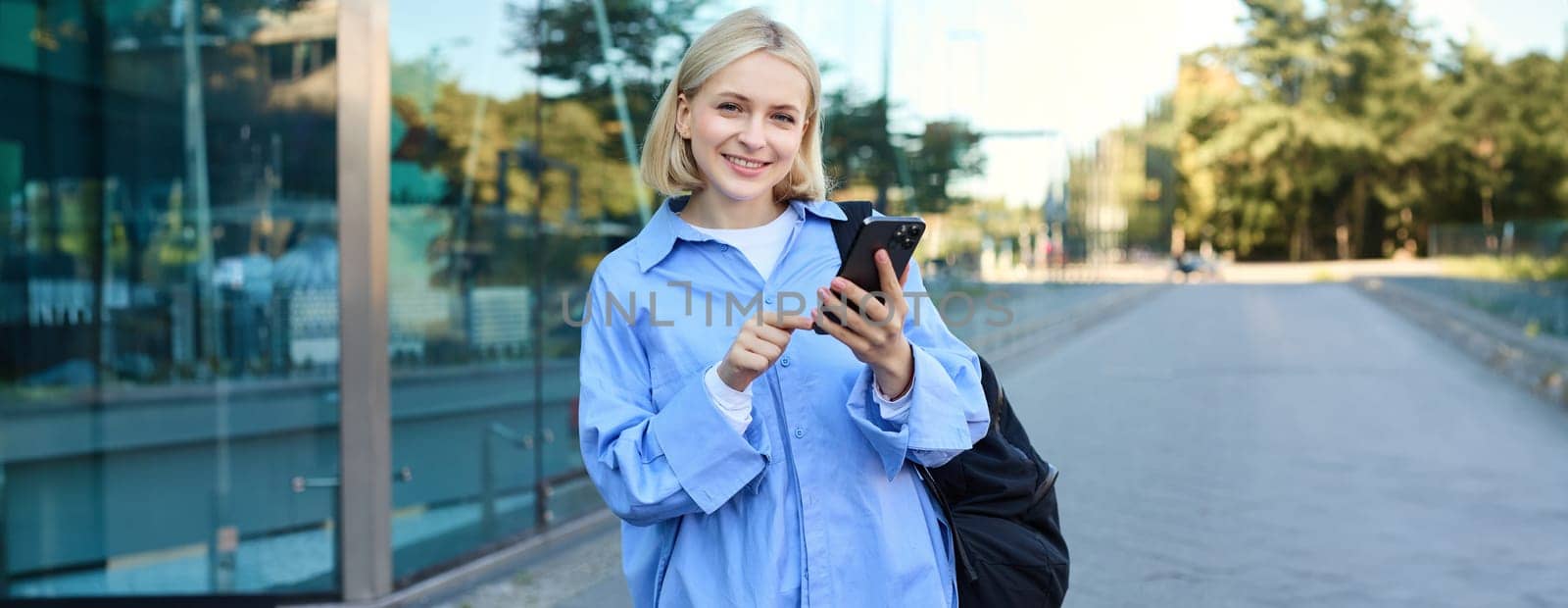 Portrait of smiling, modern blond woman walking along the street in blue collar shirt, has backpack on shoulder, messaging, using mobile phone app by Benzoix